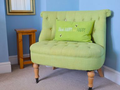 A green upholstered chair with a green cushion and a wooden side table against a blue wall at Seasides in Weymouth
