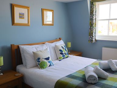 A bedroom with a wooden bed, patterned pillows, folded towels on a checkered blanket, nightstands with lamps, and framed pictures on a blue wall at Seasides in Weymouth