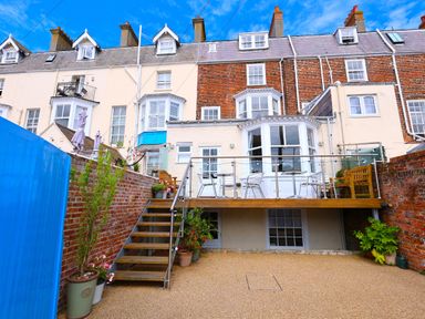 An outdoor patio area with a wooden deck, metal chairs, a wooden bench, potted plants, stairs, and multi-story brick and light-colored houses at Seasides in Weymouth