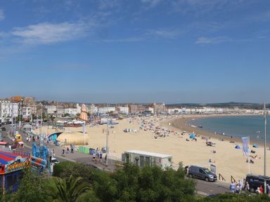 A sandy beach with people and amusement rides near a row of buildings at Seasides in Weymouth