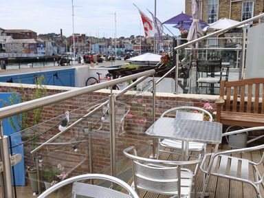 An outdoor seating area with metal chairs and table wooden bench glass railing overlooking a harbor with boats and buildings at Seasides in Weymouth
