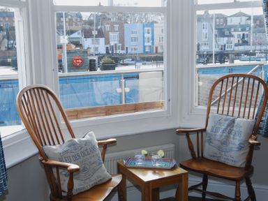 Two wooden chairs with cushions and a small wooden table with drinks in front of large windows overlooking a waterfront at Seasides Weymouth