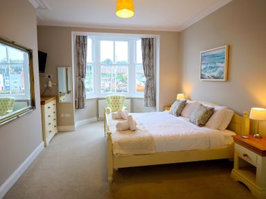 A bedroom with a bed and pillows bedside tables with lamps an armchair by the window and a chest of drawers at Seasides in Weymouth