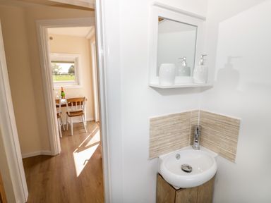 A small bathroom sink with a mirror and soap dispensers and a dining area with chairs and a bottle visible through a doorway at Malvern View Lodge in Wadborough near Pershore