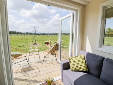 A seating area with a gray sofa and yellow cushion next to open glass doors leading to a wooden deck with two chairs and a small table overlooking a grassy field at Malvern View Lodge in Wadborough near Pershore