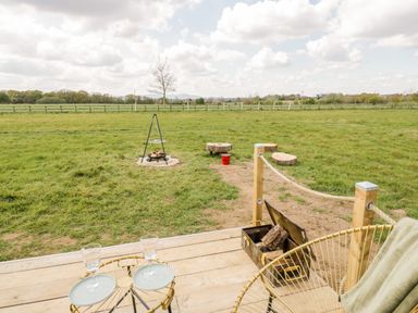 An outdoor area with wooden deck, two chairs, a table with two glasses, a fire pit, wood logs in a metal box, and tree stumps around a grassy field at Malvern View Lodge in Wadborough near Pershore