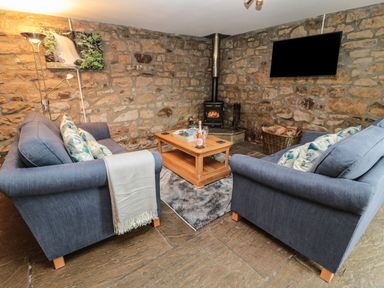 A living room with stone walls two blue sofas a wooden coffee table a wood stove and a wall-mounted TV at Granary Cottage in Bellingham