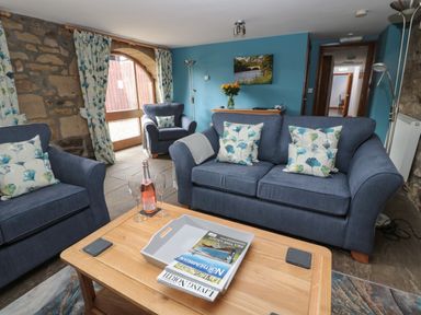 A living room with blue sofas floral cushions a wooden table with a bottle and glasses and stone walls at Granary Cottage in Bellingham