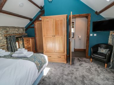 A bedroom with a bed, wooden wardrobe, dresser, black armchair, window with curtains, and an open door revealing a bathroom at Granary Cottage in Bellingham
