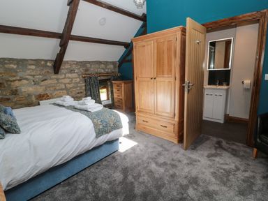 A bedroom with a bed, wooden wardrobe, chest of drawers, stone wall, and an open door leading to a bathroom at Granary Cottage in Bellingham