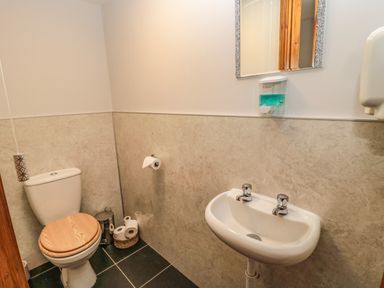 A small bathroom with a toilet with a wooden seat a sink with two taps a mirror and soap dispenser at Granary Cottage in Bellingham