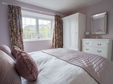 A bedroom with a bed covered with pillows and blankets a window with floral curtains a wardrobe a dresser and a mirror at Plumblands in Chapel Stile