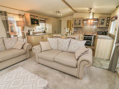 A living room with beige sofas and a patterned ottoman next to a kitchen with wooden cabinets at Lancaster Lodge in South Lakeland Leisure Village