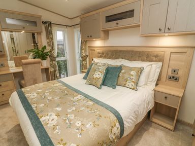 A bedroom with a bed with floral bedding and pillows a dressing table with a chair and a plant at Lancaster Lodge in South Lakeland Leisure Village
