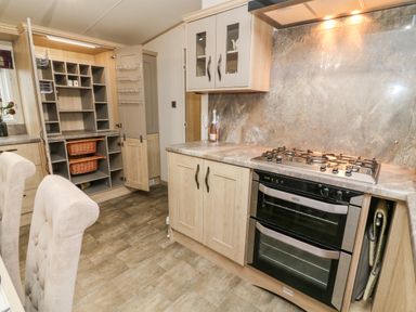 A kitchen with wooden cabinets an oven a gas stove wine bottles an open pantry and beige dining chairs at Lancaster Lodge in South Lakeland Leisure Village
