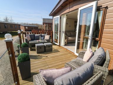 A wooden deck with wicker furniture and cushions outside a lodge with an open door at Lancaster Lodge South Lakeland Leisure Village