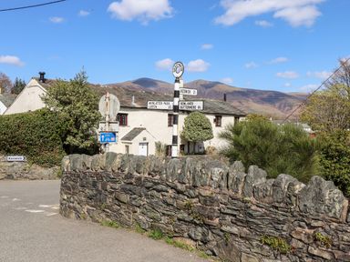 A stone wall and road signs with a white house and hills in the background at Holme Rigg in Braithwaite
