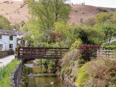A small wooden bridge over a stream with houses trees and hills in the background at Holme Rigg in Braithwaite