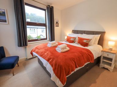 A bedroom with a double bed covered with a red blanket and white towels on top near a window with dark curtains and a chair Holme Rigg Braithwaite