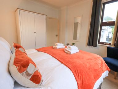 A bedroom with a bed covered with an orange blanket and pillows two white towels on the bed a white wardrobe a mirror on the wall a window with dark curtains and a blue chair at Holme Rigg in Braithwaite