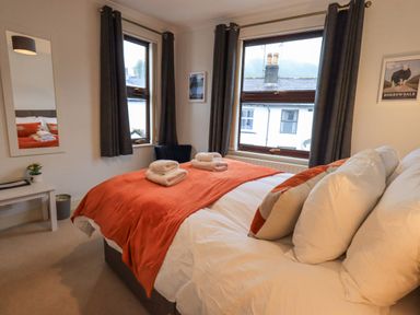 A bedroom with a bed with orange and white bedding towels on the bed two windows with dark curtains a small table with a plant and mirror on the wall at Holme Rigg in Braithwaite