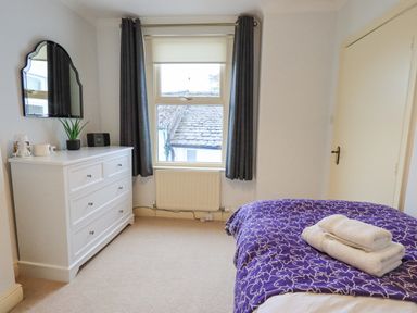 A bedroom with a bed covered in a purple patterned blanket and folded towels on top a white dresser with a mirror and items on it and a window with dark curtains at Holme Rigg in Braithwaite