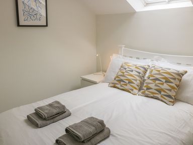 A bedroom with a white bed, patterned pillows, folded towels on the bed, a nightstand with a lamp, and a framed picture on the wall at Fig Cottage in Kirkby Stephen