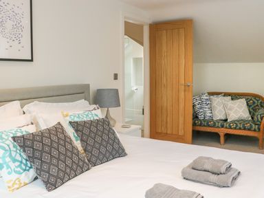 A bedroom with a bed, cushions, a side table with a lamp, an open wooden door, and a seating area with patterned cushions at Fig Cottage in Kirkby Stephen