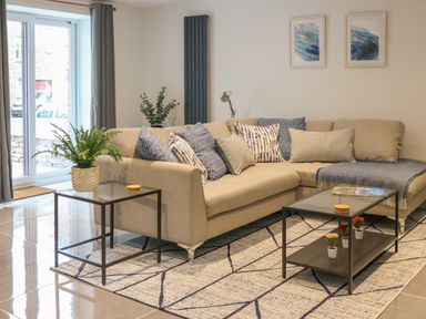 A living room with a beige sectional sofa with patterned cushions a glass coffee table with small potted flowers a side table with a plant sliding glass doors and two framed artworks on the wall at Fig Cottage in Kirkby Stephen
