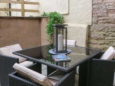 An outdoor seating area with a glass table surrounded by four cushioned wicker chairs with a candle lantern and a book on the table at Fig Cottage in Kirkby Stephen