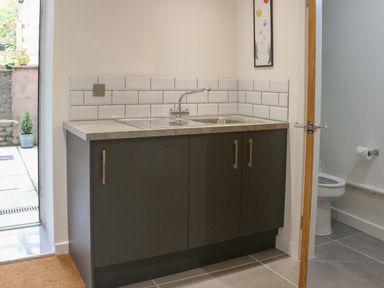 A kitchen sink with dark cabinets and white tiled backsplash next to a door leading to a bathroom with a toilet at Fig Cottage in Kirkby Stephen