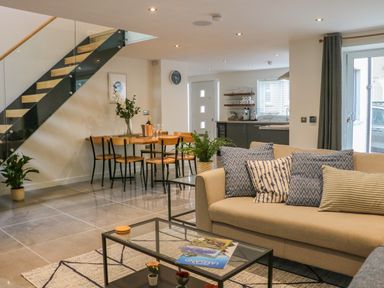 An open living and dining area with a beige sofa with patterned cushions a glass coffee table with books a wooden dining table with chairs and a staircase with glass railing at Fig Cottage in Kirkby Stephen