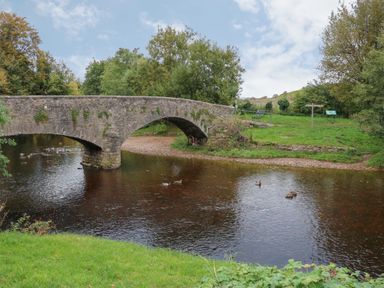 A stone arch bridge over a river with ducks swimming and grassy banks with trees at Fig Cottage in Kirkby Stephen