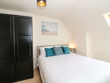 A bedroom with a bed with white bedding and teal cushions a black wardrobe a bedside table with a lamp and a beach-themed painting on the wall at Harbour Reach 6 in Weymouth
