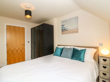 A bedroom with a bed with teal and blue pillows a wooden wardrobe a wooden door and a bedside table with a lamp at Harbour Reach 6 in Weymouth