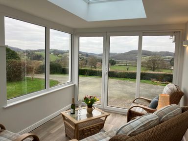 A sitting area with wicker chairs a table with flowers and a bottle of champagne by large windows overlooking hills at Berwyn View in Trelydan near Welshpool