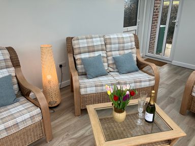 A sitting room with wicker armchairs and a loveseat with plaid cushions a floor lamp and a glass top coffee table with flowers and a bottle at Berwyn View in Trelydan near Welshpool