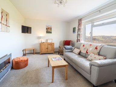A living room with a grey sofa a wooden coffee table an armchair a wall-mounted TV and a window showing a rural view at Berwyn View Trelydan near Welshpool