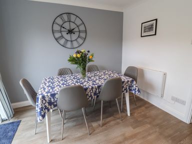 A dining room with a table covered by a blue and white tablecloth surrounded by six gray chairs and a large wall clock at Beach Cottage in Portreath