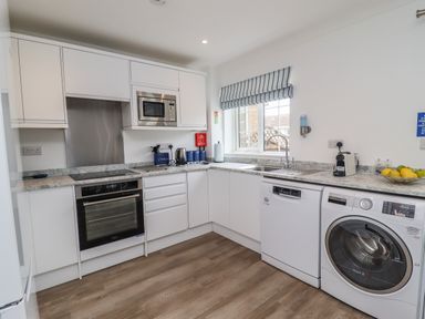 A kitchen with white cabinets oven microwave dishwasher washing machine and a window with striped blind at Beach Cottage in Portreath