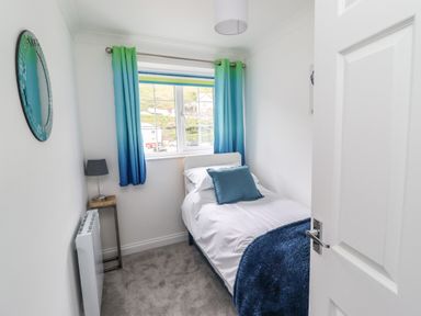 A small bedroom with a single bed a blue pillow blue and green curtains and a lamp on a side table at Beach Cottage in Portreath