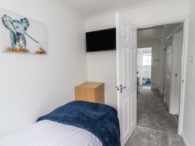 A small bedroom with a single bed a blue blanket a wooden drawer a wall-mounted TV and a door leading to a hallway and bathroom at Beach Cottage in Portreath