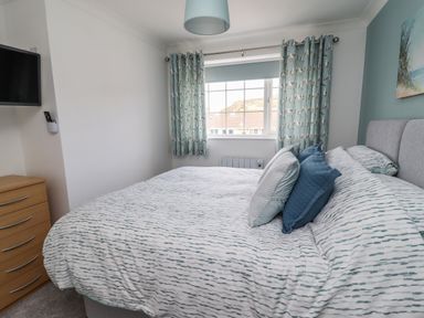 A bedroom with a bed with patterned bedding and pillows a window with patterned curtains a wooden chest of drawers and a wall-mounted television at Beach Cottage in Portreath