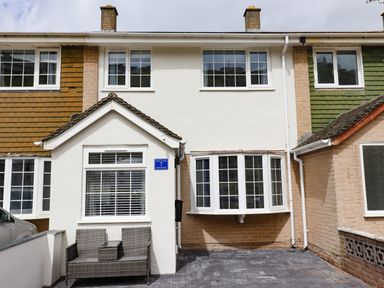 The front exterior of a two-story house with a bay window and a small porch with wicker chairs at Beach Cottage in Portreath
