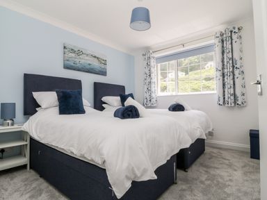 A bedroom with two single beds white bedding blue headboards and cushions a window with floral curtains and a painting on the wall at Beach Cottage in Portreath