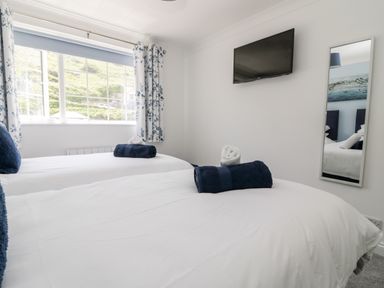A bedroom with two beds each with pillows and towels a wall mounted TV a window with curtains and a mirror at Beach Cottage in Portreath
