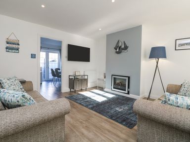 A living room with two sofas cushions a fireplace floor lamp a wall mounted tv and a dining area visible through a doorway at Beach Cottage in Portreath