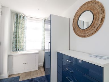 A bedroom with blue drawers a mirror on the wall and a window with curtains at Beach Cottage in Portreath