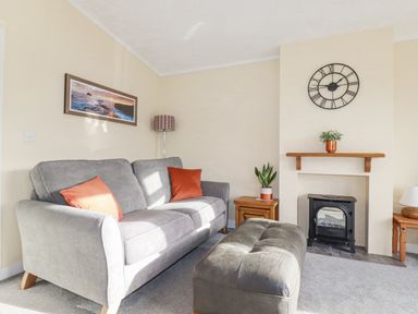 A living room with a grey sofa, orange pillows, a black stove, a wall clock, and wooden side tables at Laurel Lodge Bossiney Bay Holiday Park near Tintagel