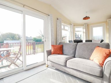 A living room with a gray sofa and orange cushions next to sliding glass doors leading to a deck with wooden chairs and table at Laurel Lodge Bossiney Bay Holiday Park near Tintagel